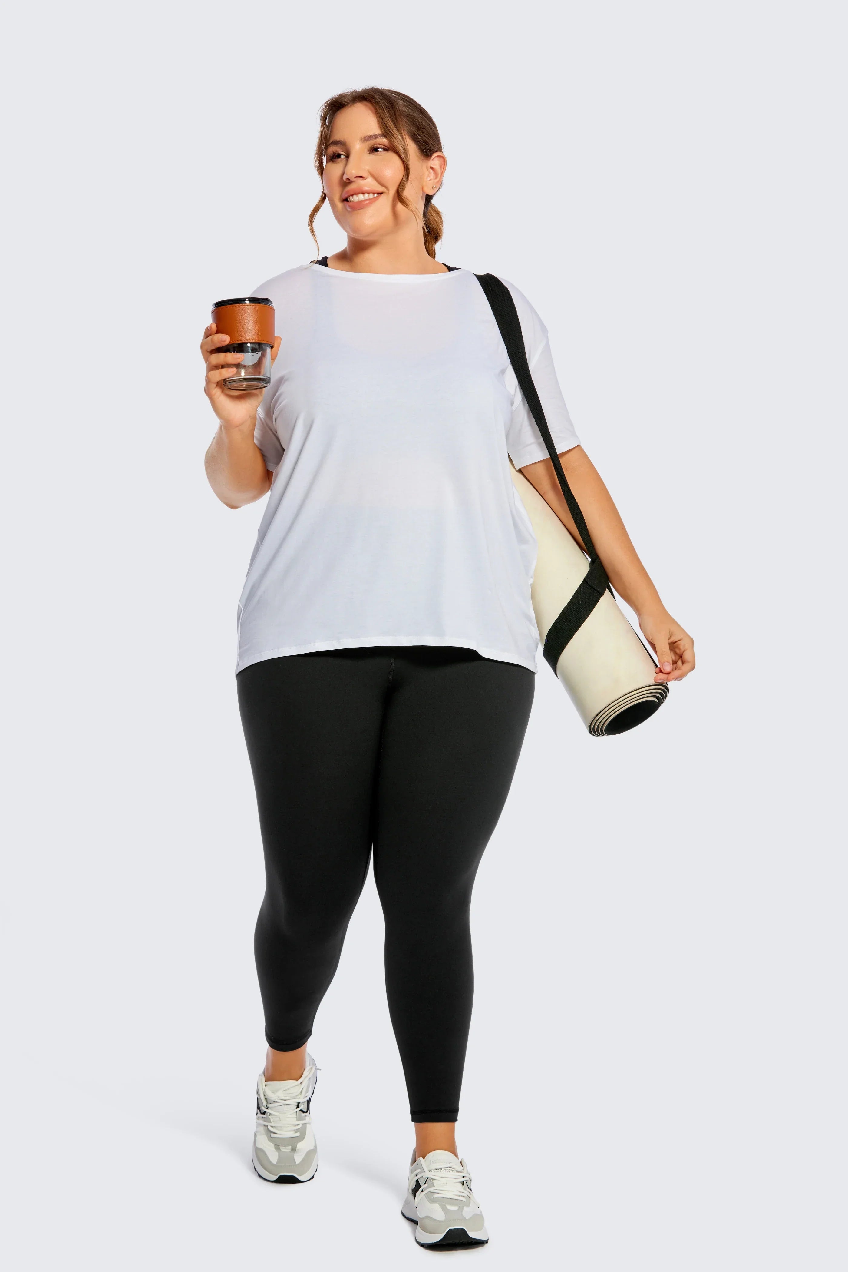 Woman wearing a white t-shirt and black leggings, holding a coffee cup and carrying a yoga mat.